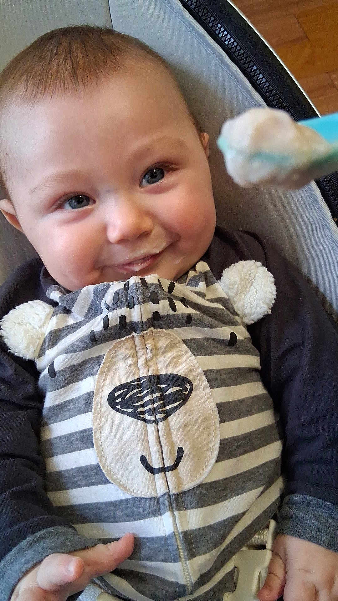 Liam joined the competition — help win amazing prizes! baby, infant, smile, milk_mustache, high_chair, feeding, spoon, food, clothing, striped_outfit, bear_face, cute, happy, child, indoor, person, closeup, hand, seat, wood_floor