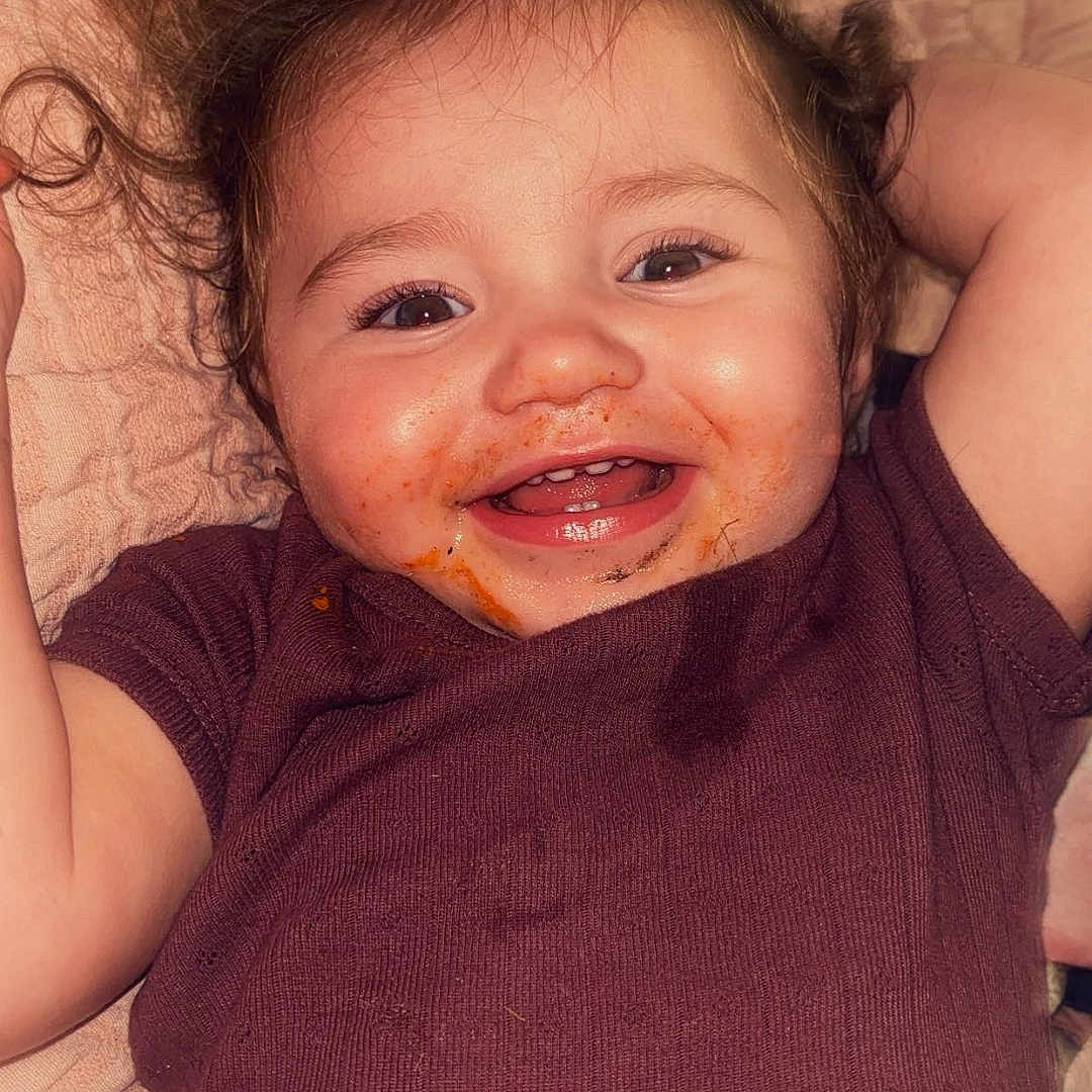 Whitley joined the competition — help win amazing prizes! baby, child, infant, smiling, happy, portrait, close_up, lying_down, blanket, bed, indoor, burgundy_clothing, dark_hair, brown_eyes, face, arms_raised, joyful, candid, cozy, snack_face