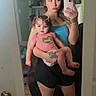 baby, infant, mother, adult, mirror_selfie, portrait, indoors, bedroom, pink_outfit, blue_top, tattoo, smartphone, mirror, devil_horns, digital_filter, sitting, standing, shelves, home_decor, wood_floor