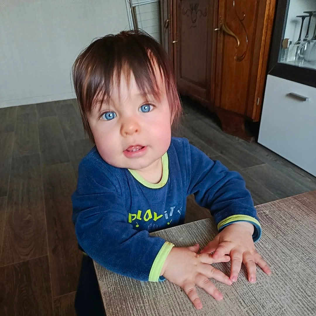 Maël participe au concours pour gagner de l'argent avec cette photo : toddler, child, blue_eyes, blue_sweater, indoor, wooden_table, hands, face, hair, floor, cabinet, furniture, portrait, person, cute, curious, young_child, home, cozy, expression