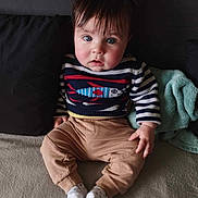 Maël participe au concours pour gagner de l'argent avec cette photo : baby, child, sitting, couch, sweater, striped_clothing, fish_pattern, pants, socks, indoor, face, hair, blue_eyes, blanket, pillow, soft_lighting, curious_expression, toddler, cozy, portrait