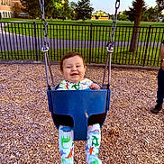 Phoenix is registered to the contest to win money with this photo: baby, child, clothing, dinosaur_pajamas, evening, fence, footwear, grass, happy, metal_chains, nature, outdoor, park, person, playground, seat, smiling, swing, trees, wood_chips