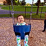 baby, swing, playground, wood_chips, fence, trees, grass, dinosaur_pajamas, child, outdoor, smiling, happy, person, evening, park, clothing, footwear, seat, metal_chains, nature