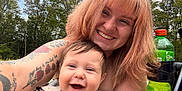 Phoenix is registered to the contest to win money with this photo: baby, bench, casual_clothing, daylight, face, family, green_bottle, hair, happy, holding, laughing, nature, outdoor, person, portrait, skin, smiling, tattoos, trees, woman