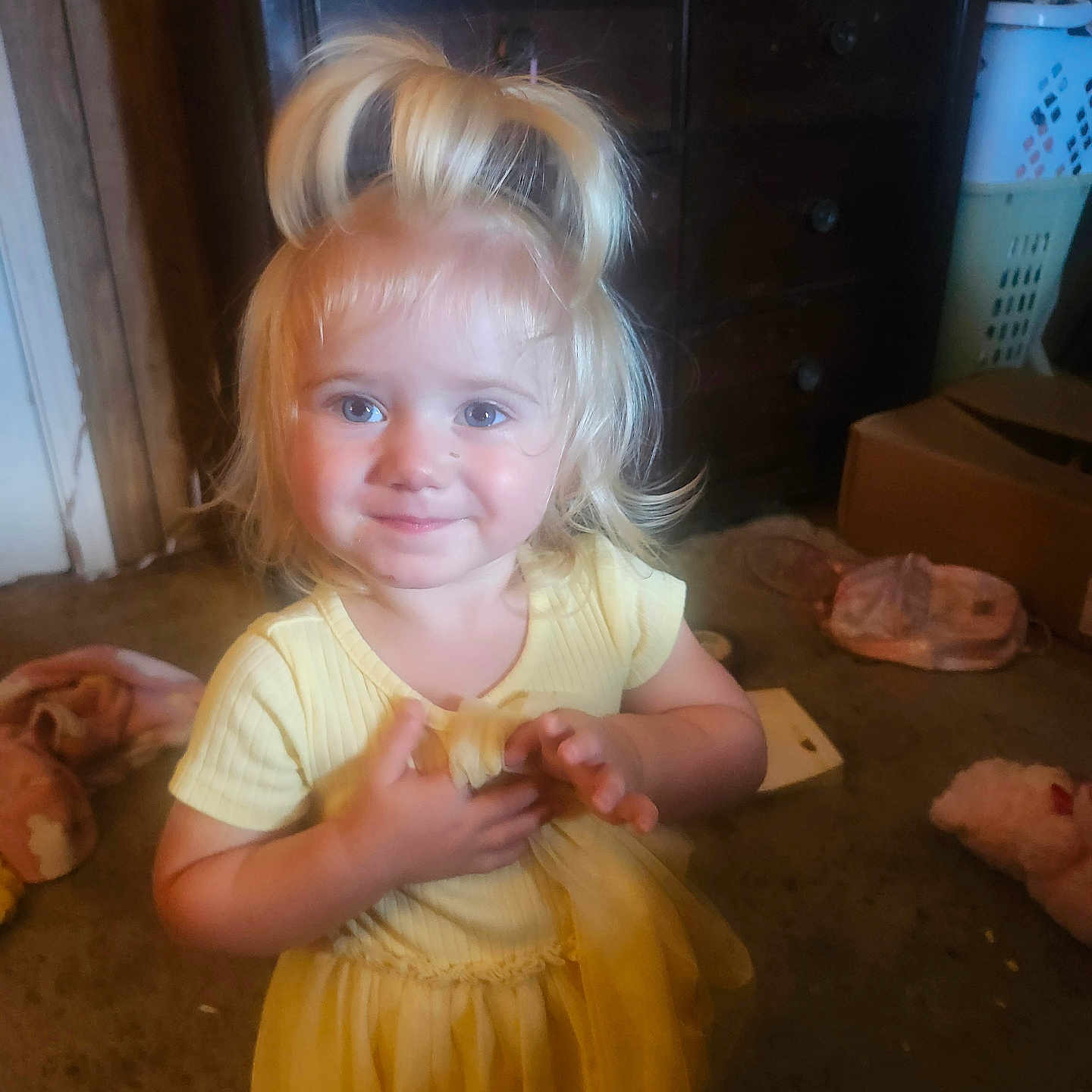 Dilylah is registered to the contest to win money with this photo: child, toddler, girl, blonde_hair, blue_eyes, smiling, yellow_dress, portrait, indoor, home, living_room, furniture, dresser, laundry_basket, shoes, cardboard_box, plush_toy, carpet, sitting, happy