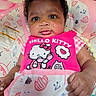 baby, infant, child, portrait, lying_down, wide_eyed, pink_clothing, hello_kitty, bib, blanket, pillow, bed, nursery, indoor, adult_hand, patterned_fabric, cute, photograph, close_up, smiling