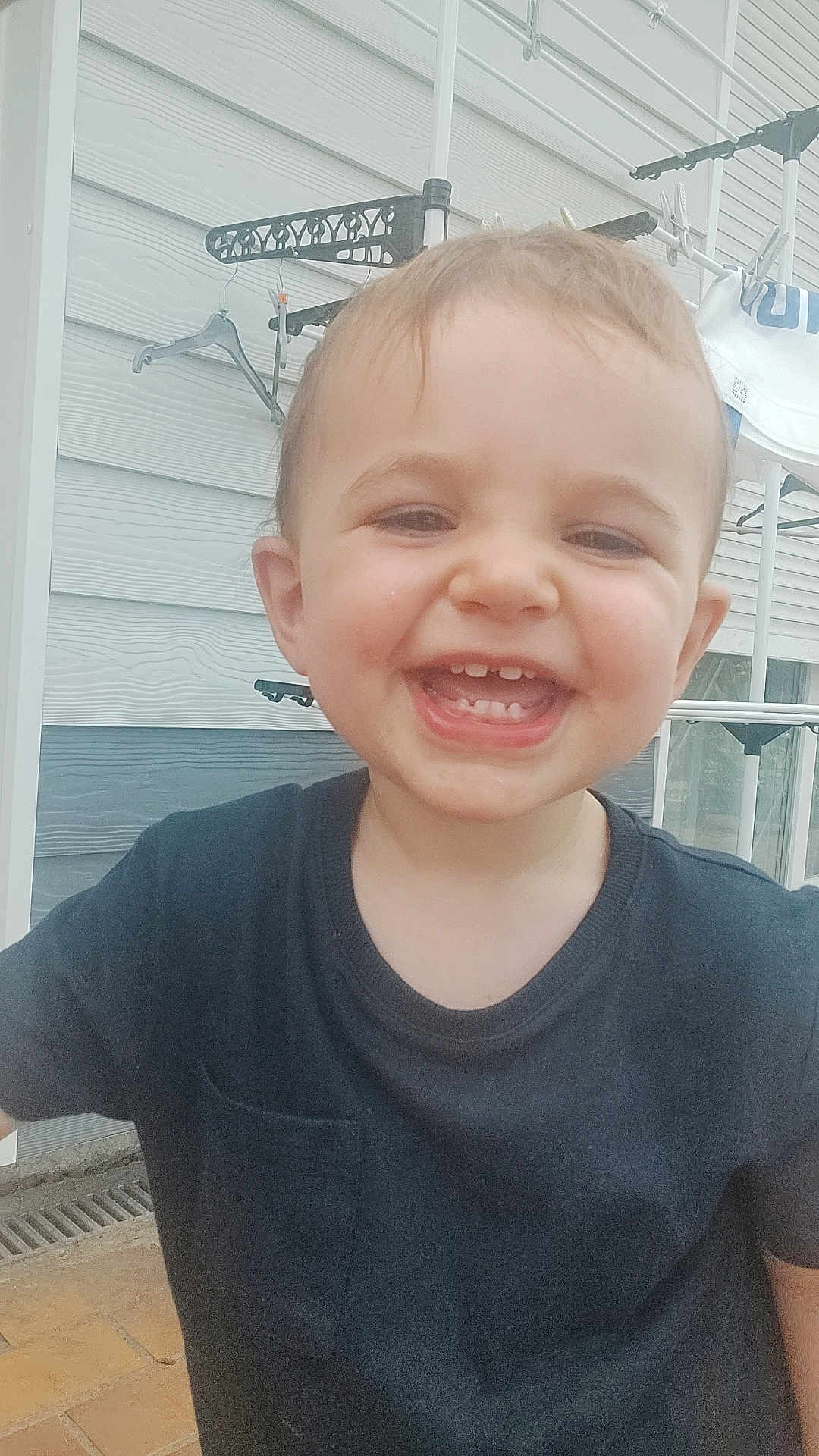 Jules participe au concours pour gagner de l'argent avec cette photo : child, toddler, smiling, black_shirt, close_up, outdoor, house_wall, hanger, drying_rack, window, happy, teeth, face, person, young_child, casual_clothing, daylight, skin, head, expression