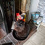 cat, black_cat, fluffy, yellow_eyes, fireplace, brick_hearth, container, paint_cans, tools, lighter, floor, tile_floor, indoor, pet, animal, feline, cozy, cluttered, home, looking_up