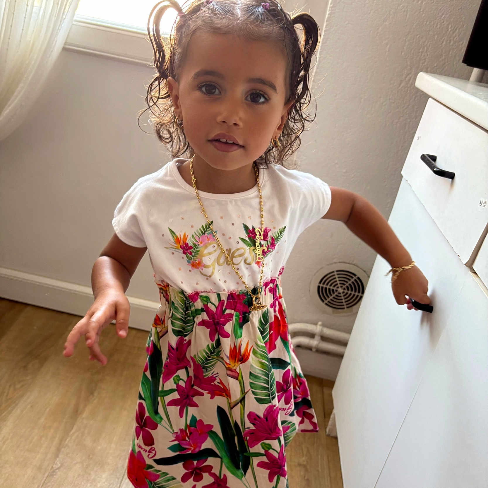Tinaya participe au concours pour gagner de l'argent avec cette photo : cabinet, child, curious_expression, curly_hair, cute, dress, floral_pattern, girl, gold_earrings, gold_necklace, indoor, jewelry, natural_light, person, pigtails, portrait, standing, white_wall, window, wooden_floor