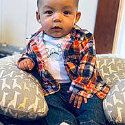 Thiago is registered to the contest to win money with this photo: baby, child, indoor, plaid_shirt, dinosaur_tee, giraffe_pillow, cushion, denim_jeans, socks, cute, portrait, seated, expression, hands, face, eyes, wooden_door, fabric, cozy, casual_clothing