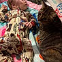 baby, cat, onesie, cowboy_theme, play_mat, tabby_cat, curious, indoor, colorful, infant, pet, animal, child, patterned_clothing, soft_texture, lying_down, looking_sideways, cute, domestic, friendly