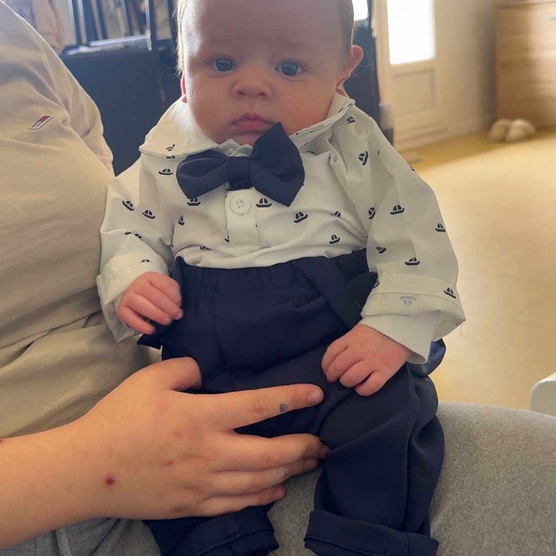 Naylan participe au concours pour gagner de l'argent avec cette photo : baby, bow_tie, shirt, pants, barefoot, person, hand, indoor, furniture, chair, door, curious, serious_expression, seated, casual_clothing, floor, natural_light, adult_hand, infant, portrait