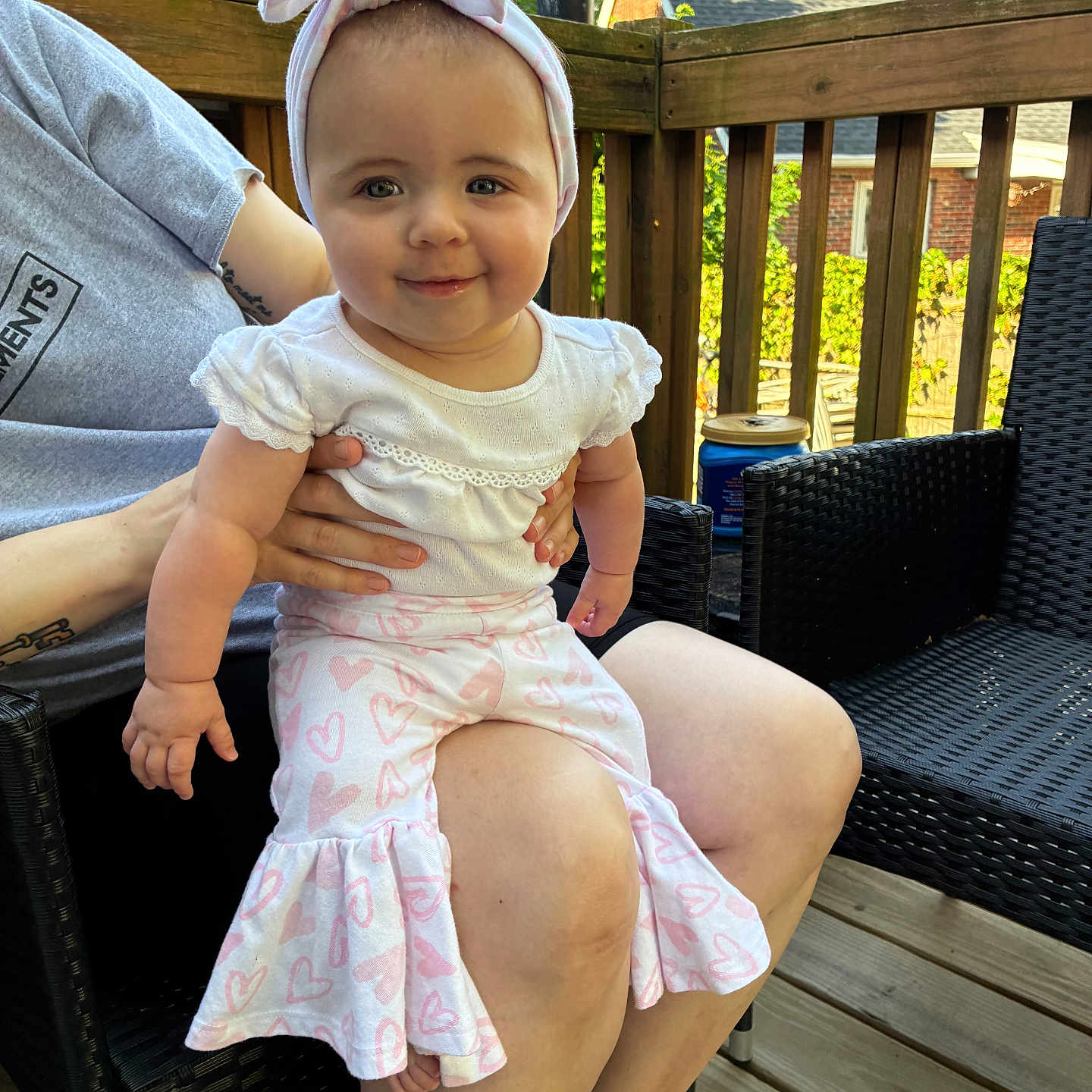 Chrissy is registered to the contest to win money with this photo: baby, casual_clothing, chair, child, daylight, furniture, happy, headband, heart_pattern, human_hand, infant, lap, outdoor, pants, person, portrait, skin, smiling, white_shirt, wooden_deck