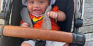Kamari is registered to the contest to win money with this photo: baby, child, stroller, hat, bucket_hat, smiling, happy, bracelet, orange_shirt, shorts, barefoot, outdoor, wooden_deck, seatbelt, black_seat, leather_handlebar, person, infant, cute, sun_hat
