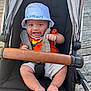 baby, child, stroller, hat, bucket_hat, smiling, happy, bracelet, orange_shirt, shorts, barefoot, outdoor, wooden_deck, seatbelt, black_seat, leather_handlebar, person, infant, cute, sun_hat