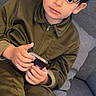 child, boy, olive_green_clothing, smartphone, sitting, couch, gray_couch, indoors, curious, young, portrait, casual, holding, technology, home, furniture, person, face, hair, expression