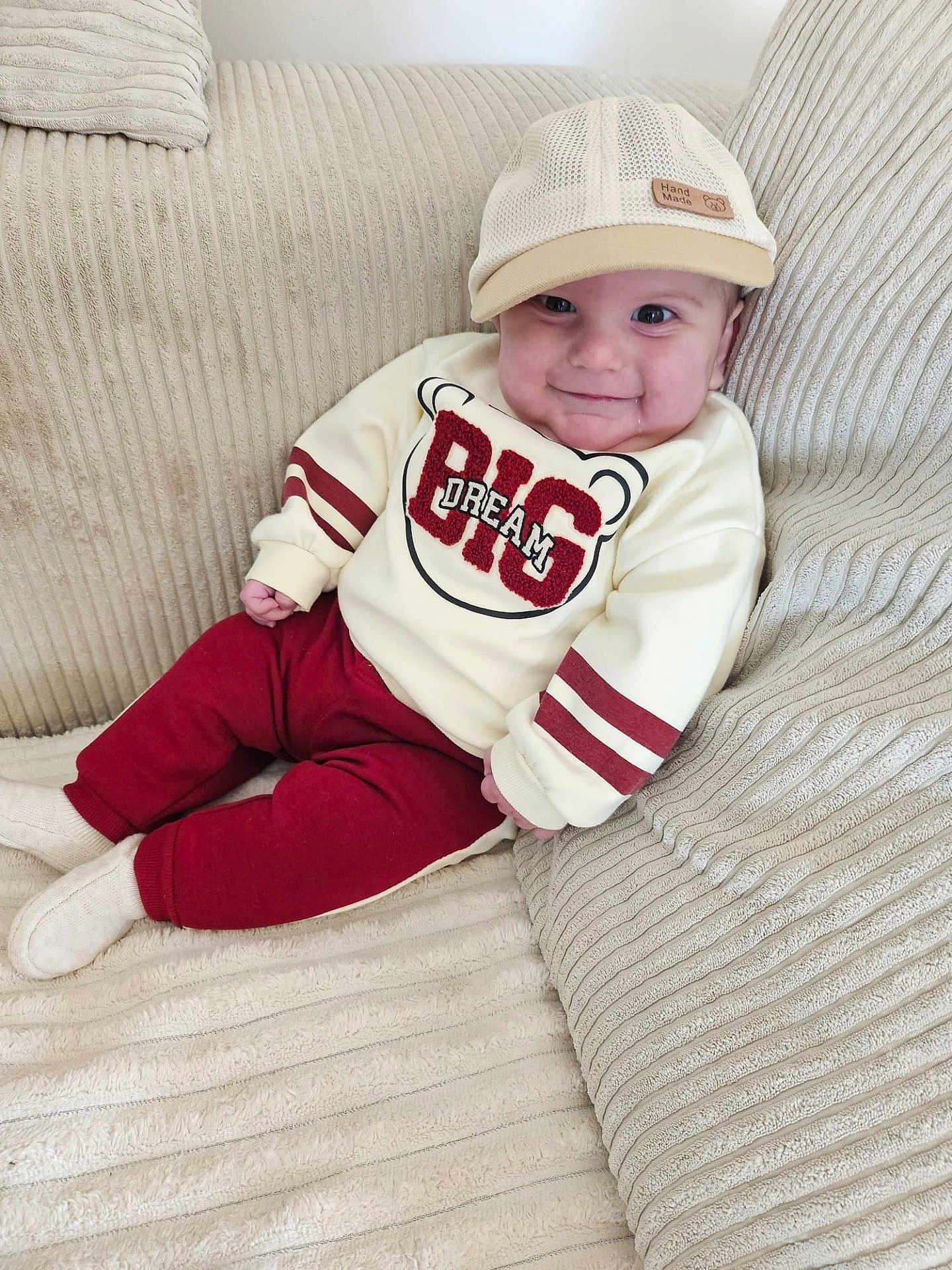 Andrea a rejoint le concours — aidez-le/la à gagner de superbes lots ! baby, infant, smiling, hat, cap, sweatshirt, text_on_clothing, pants, socks, sofa, cushion, beige, striped_texture, cozy, portrait, cute, cheeks, handmade_label, drool, sitting