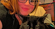 Jake is registered to the contest to win money with this photo: person, cat, black_cat, glasses, indoor, blanket, bedroom, smile, relaxing, wall_art, butterflies, shelf, furniture, cozy, pet, cute, whiskers, hair, casual, resting