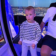 William a rejoint le concours — aidez-le/la à gagner de superbes lots ! child, boy, striped_sweater, pants, indoor, blue_lighting, cityscape, window, night, adult, shorts, seat, ferris_wheel, observation_pod, expression, curious, person, casual_clothing, hand, leg