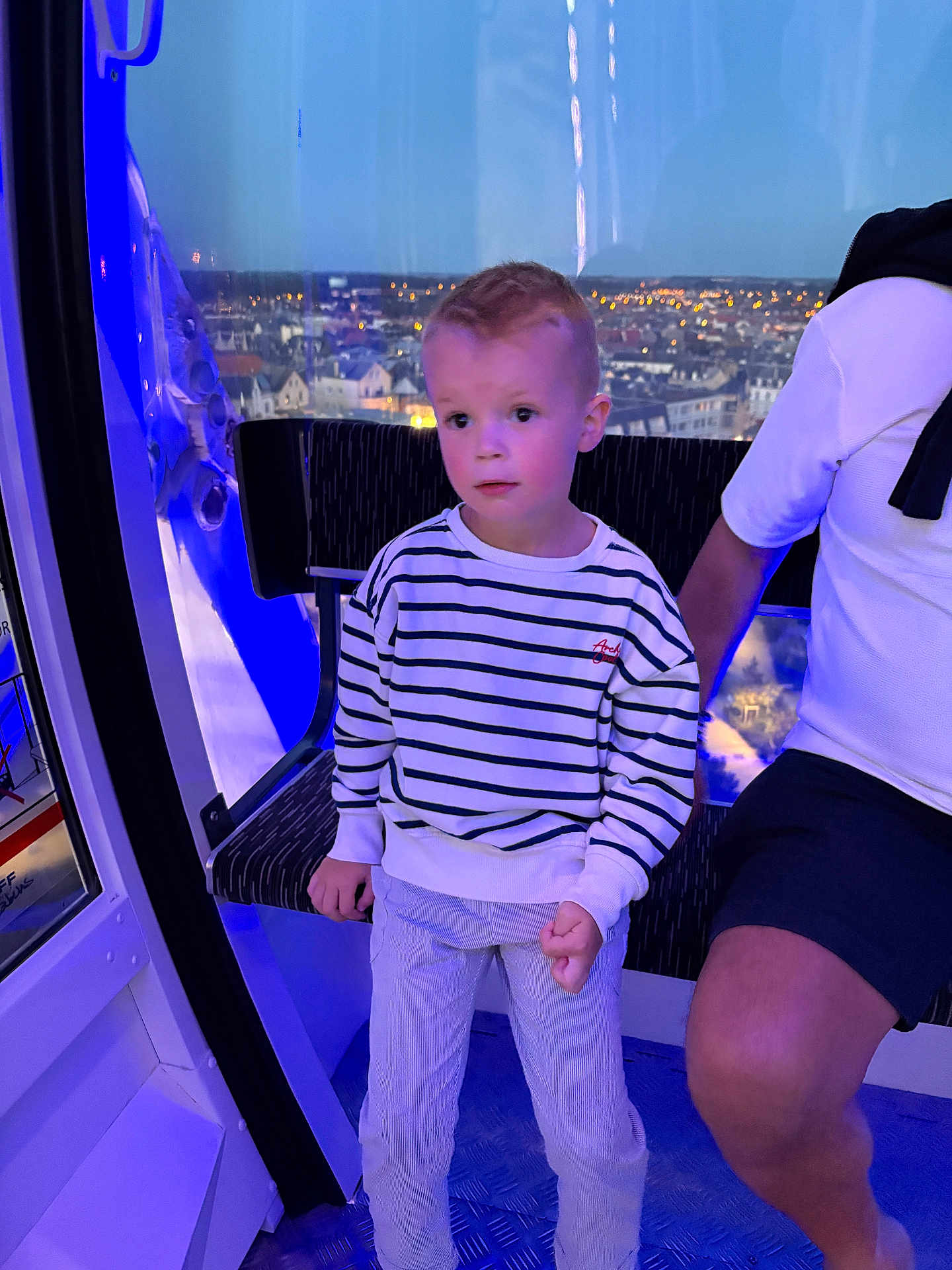 William a rejoint le concours — aidez-le/la à gagner de superbes lots ! child, boy, striped_sweater, pants, indoor, blue_lighting, cityscape, window, night, adult, shorts, seat, ferris_wheel, observation_pod, expression, curious, person, casual_clothing, hand, leg