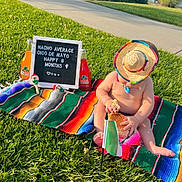 Angelo is registered to the contest to win money with this photo: baby, sombrero, sandwich, mexican_flag, blanket, grass, maracas, jarritos, bottle, letter_board, outdoor, sunny, child, celebration, colorful, picnic, food, sitting, summer, cute