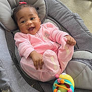 Malani is registered to the contest to win money with this photo: baby, child, infant, smiling, happy, portrait, sitting, rocker, bouncer, pink_outfit, hair_bow, toy, colorful_toy, indoor, living_room, carpet, furniture, face, playtime, joyful