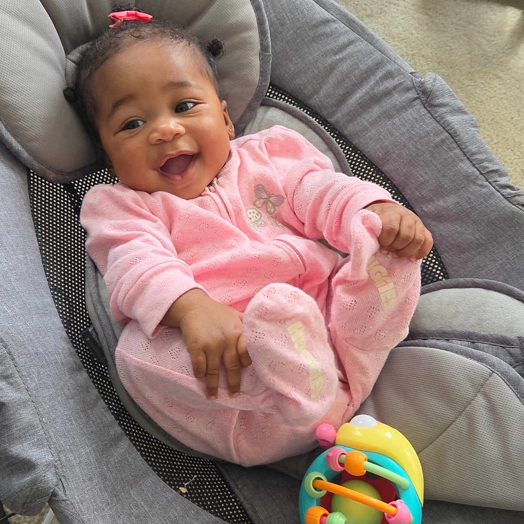Malani is registered to the contest to win money with this photo: baby, child, infant, smiling, happy, portrait, sitting, rocker, bouncer, pink_outfit, hair_bow, toy, colorful_toy, indoor, living_room, carpet, furniture, face, playtime, joyful