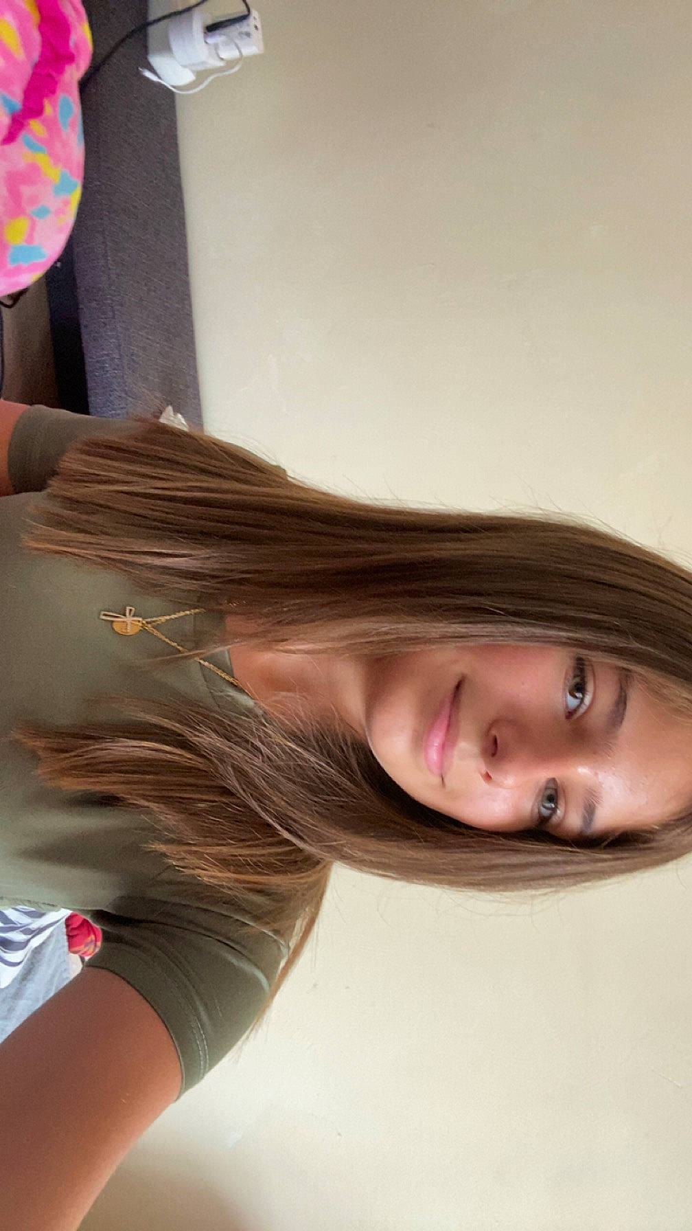 accessory, casual, cross_pendant, face, female, green_shirt, hair, indoor, jewelry, long_hair, natural_light, necklace, person, portrait, relaxed, shoulder, skin, smile, wall, young