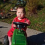 toddler, child, toy, tractor, outdoor, red_clothing, steering_wheel, grass, sidewalk, sunlight, shadow, green_toy, play, serious_expression, nature, autumn, leaf, fun, young_child, recreation