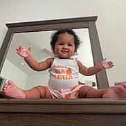 Aniya is registered to the contest to win money with this photo: baby, infant, smiling, mirror, wooden_furniture, arms_outstretched, cute_clothing, indoors, child, happy, sitting, face, curly_hair, room, reflection, white_wall, foot, portrait, person, casual