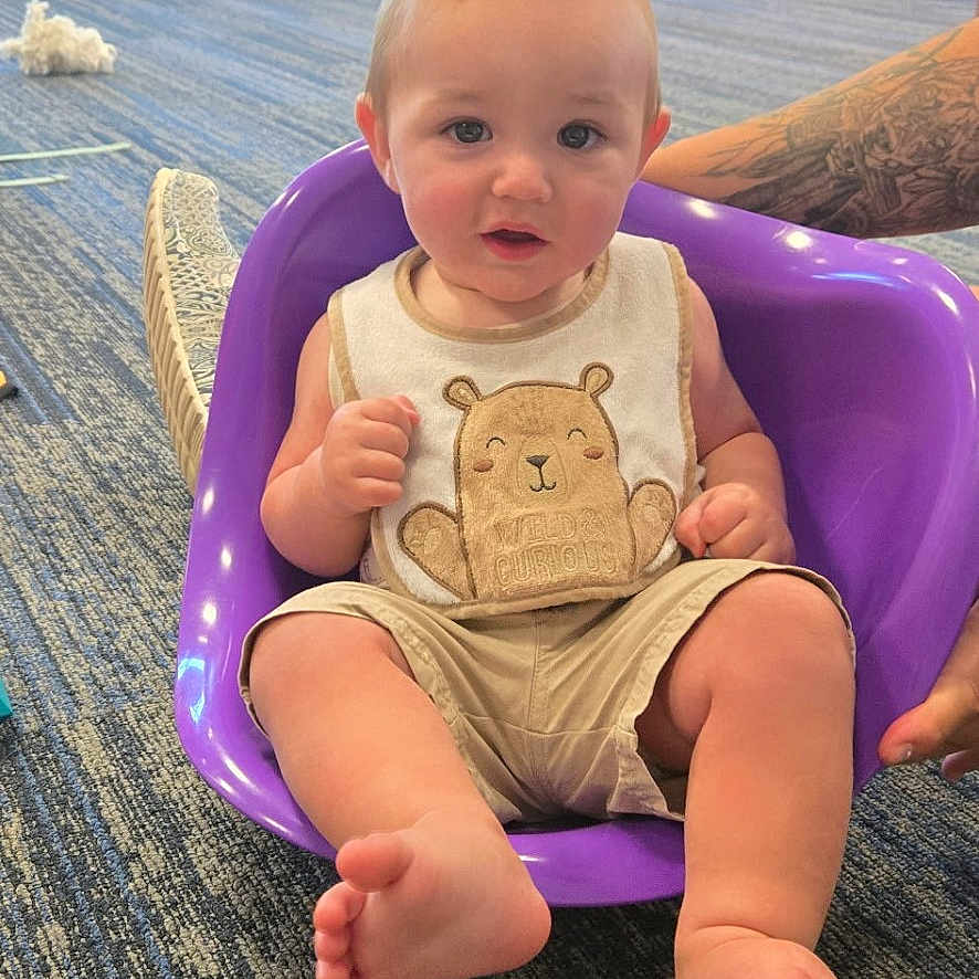 Pj is registered to the contest to win money with this photo: baby, child, indoor, purple_seat, carpet, bear_design, clothing, person, floor, play_tunnel, window, daylight, tattoo, foot, hand, child_playing, chair, background, colorful, happy