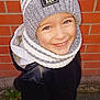 child, smiling, hat, pom_pom, scarf, jacket, sneakers, brick_wall, outdoor, happy, blue_eyes, winter_clothing, street, casual_wear, portrait, person, standing, fashion, warm_clothes, daylight