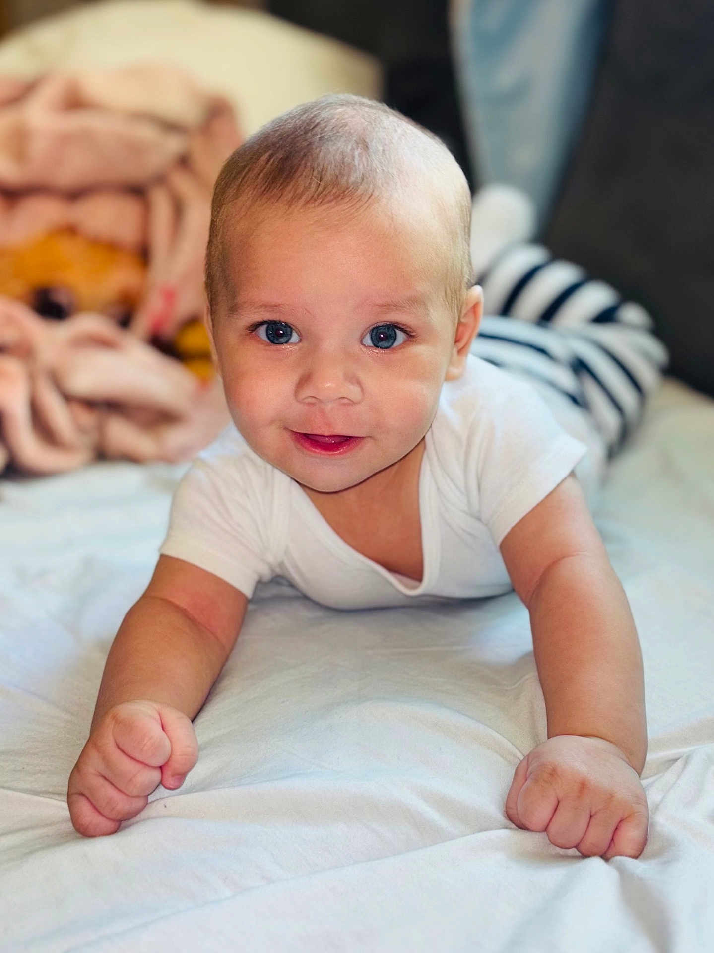 Jaidyn is registered to the contest to win money with this photo: baby, infant, smiling, face, blue_eyes, white_shirt, striped_pants, bed, blanket, cute, child, skin, portrait, hands, lying_down, indoors, soft_light, happy, toddler, closeup