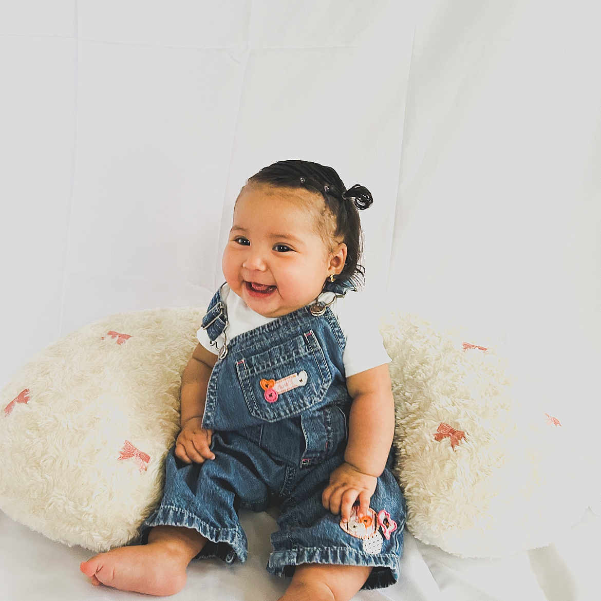 Korayma is registered to the contest to win money with this photo: adorable, baby, barefoot, child, cushion, cute, denim_overalls, hair_tie, happy, indoors, infant, pink_bows, portrait, sitting, small_earrings, smile, soft_texture, studio, white_background, white_shirt