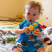 Richard is registered to the contest to win money with this photo: toddler, child, toy, blue_clothing, mickey_mouse, indoor, blanket, stuffed_animal, curly_hair, face, playtime, colorful_toy, expression, sitting, soft_toys, bedroom, cute, young_child, person, home