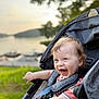 baby, smiling, stroller, child, outdoor, nature, grass, water, lake, trees, happy, pointing, daylight, person, face, blue_eyes, clothing, seatbelt, photography, infant