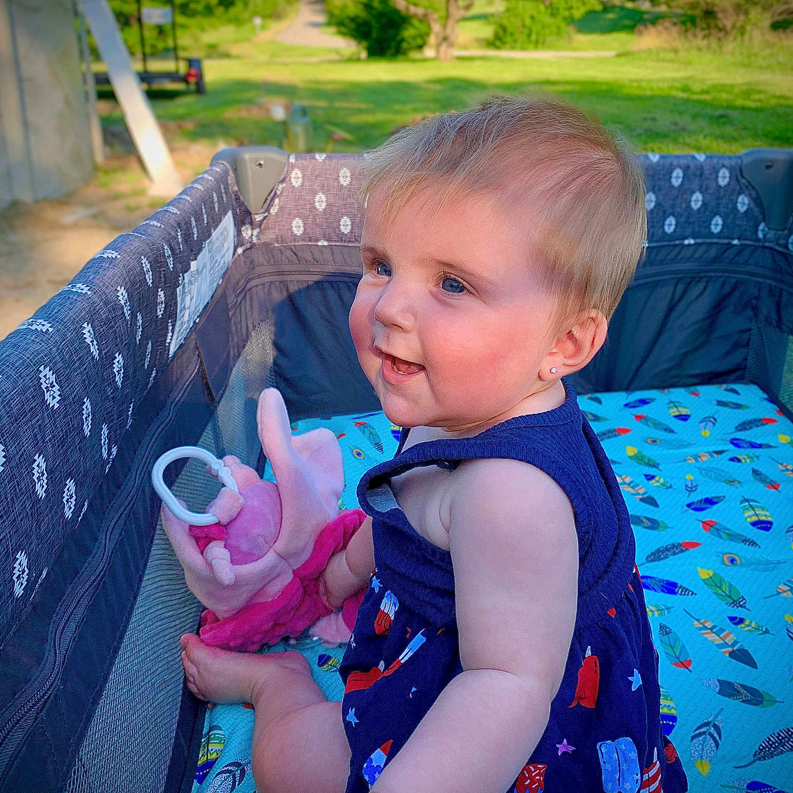 Kynzlie is registered to the contest to win money with this photo: baby, blue_dress, child, cute, face, grass, greenery, happy, infant, outdoor, park, playpen, portrait, sitting, skin, smiling, soft_toy, summer, sunlight, toy