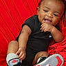 baby, infant, smiling, black_clothing, sneakers, red_background, happy, child, portrait, cute, sitting, hand, footwear, young_child, indoors, person, fashion, casual, playful, closeup