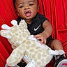 Zaccai is registered to the contest to win money with this photo: baby, child, infant, toy, plush_giraffe, black_clothing, nike_logo, red_background, sitting, curious, open_mouth, cute, indoor, footwear, socks, portrait, young_child, soft_toy, happy, playful