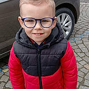 Dario participe au concours pour gagner de l'argent avec cette photo : child, boy, glasses, red_jacket, black_jacket, blue_pants, white_shoes, smiling, outdoor, car, cobblestone, street, vehicle, standing, casual_clothing, young, portrait, happy, daylight, urban