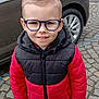 child, boy, glasses, red_jacket, black_jacket, blue_pants, white_shoes, smiling, outdoor, car, cobblestone, street, vehicle, standing, casual_clothing, young, portrait, happy, daylight, urban