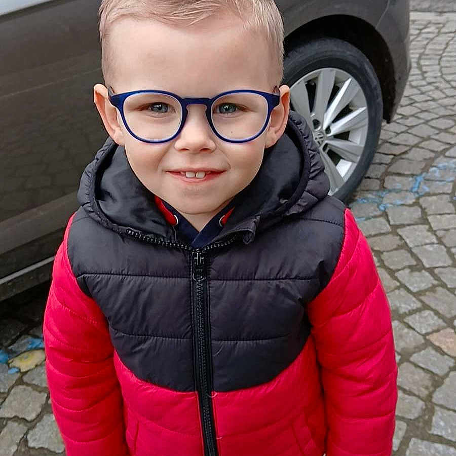 Dario participe au concours pour gagner de l'argent avec cette photo : black_jacket, blue_pants, boy, car, casual_clothing, child, cobblestone, daylight, glasses, happy, outdoor, portrait, red_jacket, smiling, standing, street, urban, vehicle, white_shoes, young