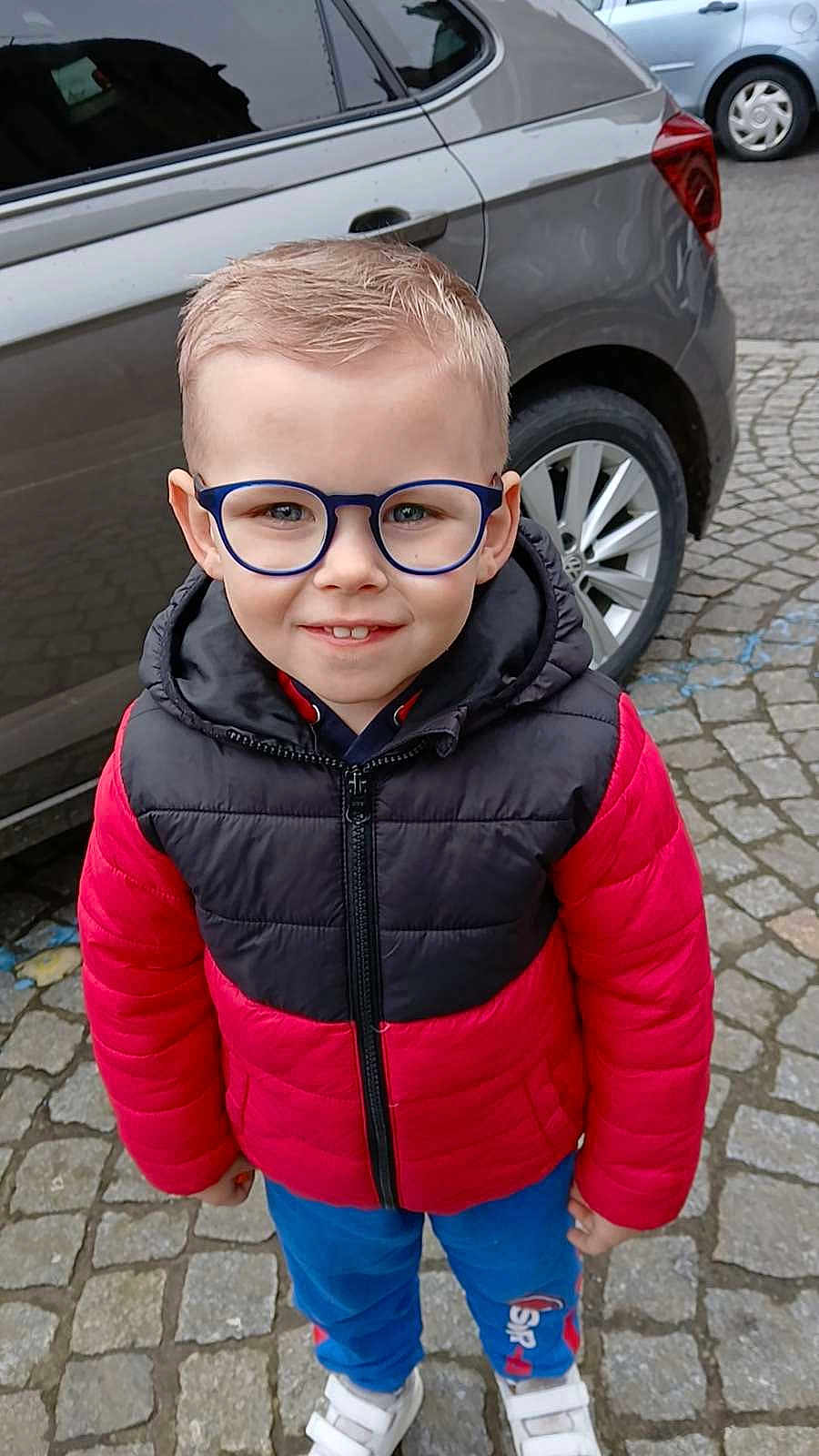 Dario participe au concours pour gagner de l'argent avec cette photo : child, boy, glasses, red_jacket, black_jacket, blue_pants, white_shoes, smiling, outdoor, car, cobblestone, street, vehicle, standing, casual_clothing, young, portrait, happy, daylight, urban