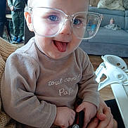 Nélya participe au concours pour gagner de l'argent avec cette photo : toddler, child, glasses, tongue_out, toy_car, indoor, living_room, person, sweater, adult_hand, couch, boxes, background, smile, playful, curly_hair, seat, furniture, holding, cozy