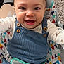 baby, smiling, child, blue_overalls, curly_hair, face, infant, seat, patterned_fabric, happy, cute, indoors, clothing, person, closeup, baby_seat, button, carpet, toddler, reaching