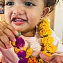 toddler, child, face, flower_garland, purple_flower, yellow_flowers, orange_flowers, paint_on_forehead, curly_hair, pink_clothing, earring, happy, close_up, indoor, cute, holding, portrait, smiling, young_child, celebration