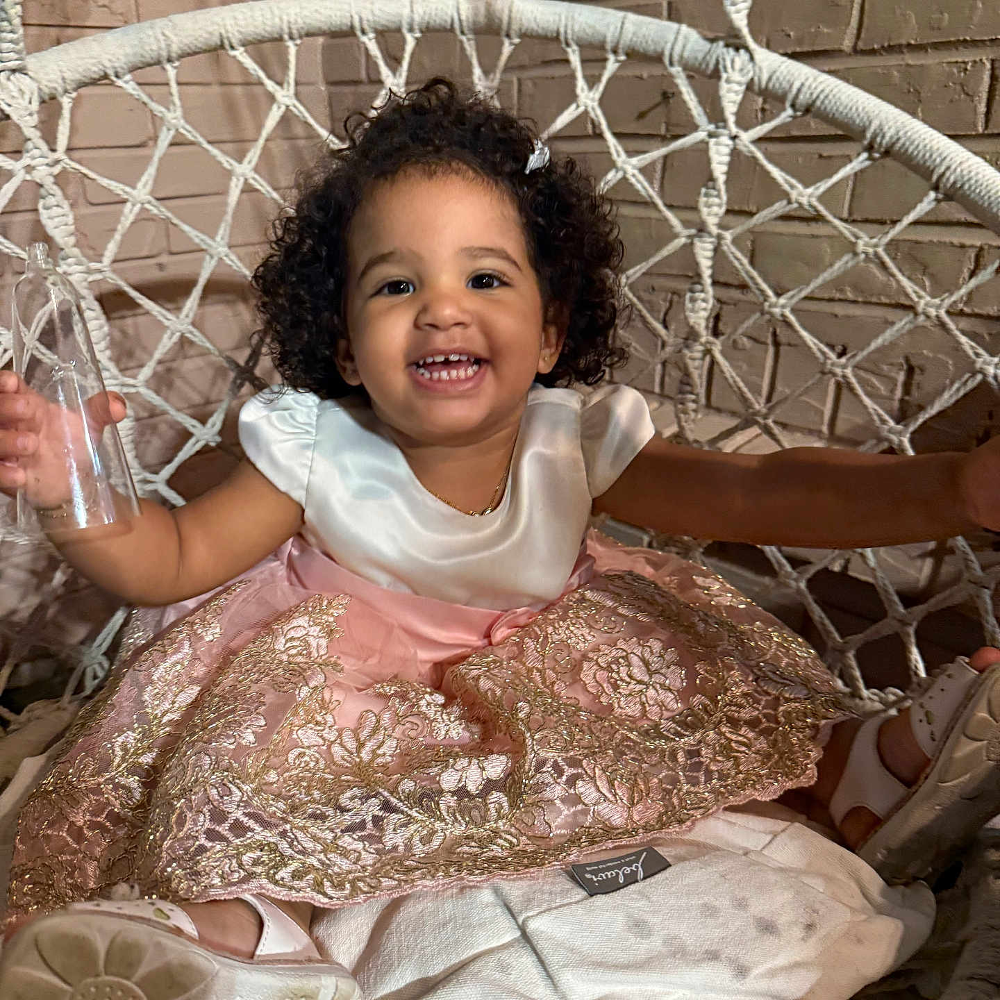 Orlansh Valdespino is registered to the contest to win money with this photo: brick_wall, child, curly_hair, cute, dress, footwear, happy, holding_object, indoors, joyful, macrame, pink_dress, plastic_bottle, portrait, sandals, seated, smiling, swing_chair, toddler, white_top