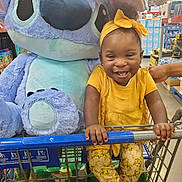 Emma is registered to the contest to win money with this photo: toddler, child, shopping_cart, plush_toy, stitch, yellow_clothing, headband, smile, grocery_store, aisle, toy, person, happy, indoor, retail, cute, holding, sneakers, fashion, fun