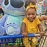 toddler, child, shopping_cart, plush_toy, stitch, yellow_clothing, headband, smile, grocery_store, aisle, toy, person, happy, indoor, retail, cute, holding, sneakers, fashion, fun