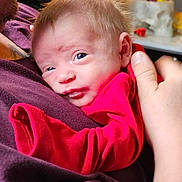 John is registered to the contest to win money with this photo: baby, infant, red_clothing, purple_shirt, adult_hand, face, eyes, skin, hair, indoor, blurred_background, close_up, snuggling, person, holding, newborn, soft_light, portrait, cute, cozy